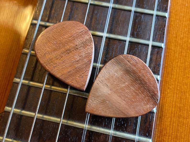 May include: Two wooden guitar picks with a teardrop shape rest on the strings and fretboard of a guitar. The picks have a reddish-brown wood grain pattern. The fretboard is dark brown, and the guitar body is a light brown.
