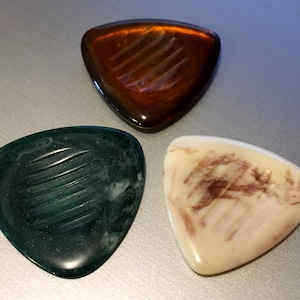 May include: Three guitar picks, one brown, one green, and one white with brown streaks. Each pick has a textured surface.
