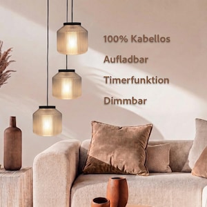 May include: Three hanging pendant lights with ribbed glass shades and black caps, suspended by thin black cords. The text on the wall reads 100% Kabellos, Aufladbar, Timerfunktion, Dimmbar, indicating the lights are cordless, rechargeable, dimmable, and have a timer function.