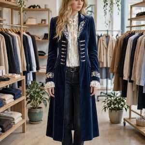 Op de afbeelding: Een lange marineblauwe fluwelen jas met zilveren knopen, wit bloemenborduursel op de revers en manchetten, en witte kanten afwerking bij de polsen. Gedragen over een witte blouse met ruches en een donkerblauwe spijkerbroek met riem.