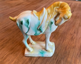 May include: A ceramic horse figurine with a yellow, white, and green glaze. The horse is standing on a square base and has a saddle.