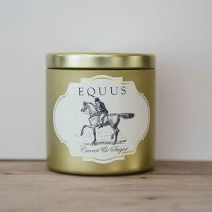 May include: A gold-colored candle tin with a label featuring a black and white illustration of a person riding a horse. The label reads "EQUUS" and "Carrot & Sugar". The candle tin is cylindrical.