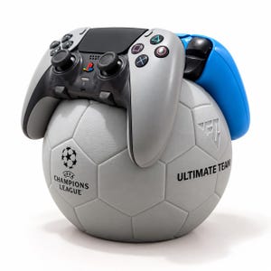 May include: A gray soccer ball stand featuring the UEFA Champions League logo and the text ULTIMATE TEAM, topped with a gray and black PlayStation 5 controller and a blue controller handle resting on the surface of the ball.