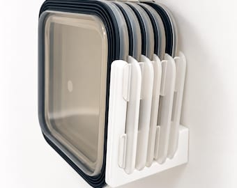 May include: A white plastic wall-mounted organizer holding a stack of square food storage container lids. The lids are translucent with dark gray rims, and the organizer features vertical slots to keep the lids upright and organized.