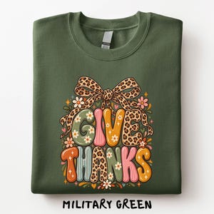 May include: A military green crewneck sweatshirt featuring a graphic print that reads GIVE THANKS in colorful, patterned bubble letters. The design includes a large leopard print bow at the top, small flower accents, and star shapes.