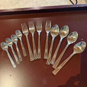 May include: A collection of twelve stainless steel utensils arranged in a fan shape, consisting of four forks and eight spoons of varying sizes, all featuring a simple, smooth handle design with a polished metallic finish.