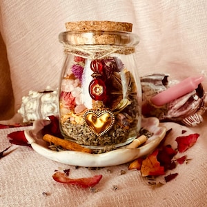 May include: A clear glass jar with a cork stopper filled with dried flowers, herbs, and small pink stones. A twine string wraps around the neck, holding a red bead charm and a gold-toned metal heart pendant. The jar sits on a small white dish.