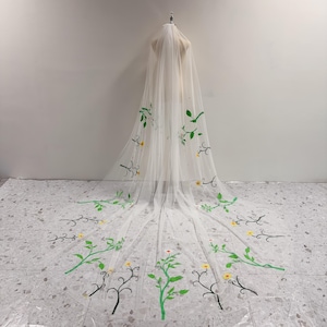 May include: A long, sheer white wedding veil featuring embroidered green vines and small yellow flowers. The delicate tulle fabric drapes downwards, showcasing a botanical pattern of winding stems and leaves that extend towards the hem.