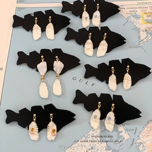 May include: Six pairs of earrings displayed on black fish-shaped cards. Each earring is made of white shell with gold accents and a small blue or white bead. The earrings are displayed on a map of Alaska.
