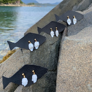 May include: Four pairs of white shell earrings with gold posts displayed on black fish-shaped cards. The cards are arranged on a rocky surface with a body of water in the background.