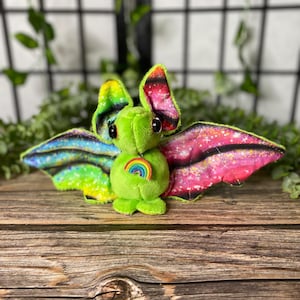 May include: A plush bat toy with a green body, rainbow wings, and a rainbow patch on its chest. The wings feature a colorful design with pink, blue, and yellow hues. The bat is set against a wooden background.