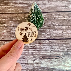 May include: A round wooden bauble with the text "Christmas Hug" and a small Christmas tree design. The bauble is held in a hand against a woodgrain background.