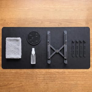 May include: A black rectangular desk mat featuring a gray microfiber cloth, a circular black coaster with a slotted pattern, a clear spray bottle, a foldable dark gray laptop stand, and four black vertical cable management clips.