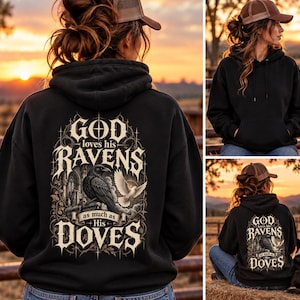 May include: A black hooded sweatshirt featuring a graphic on the back with the text "GOD loves his RAVENS as much as His DOVES." The design includes a black raven perched on stone ruins and a white dove in flight, surrounded by ornate gothic arches.