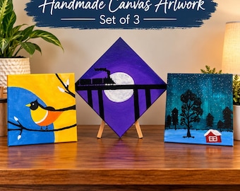 Op de afbeelding: Een set van drie handgemaakte canvas schilderijen. Het linker vierkante canvas toont een vogel op een tak tegen een gele en blauwe achtergrond. Het middelste ruitvormige canvas toont een trein op een brug voor een volle maan. Het rechter vierkante canvas toont een besneeuwd landschap met bomen en een klein rood huisje.