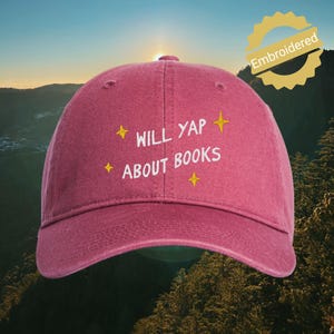 May include: A dark pink baseball cap featuring white embroidered text that reads WILL YAP ABOUT BOOKS, accented by four small yellow embroidered star shapes. The cap has a structured crown and a curved brim, with a classic six-panel design.