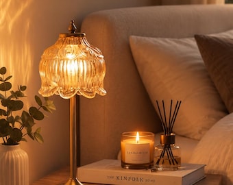 Peut inclure: Une lampe de table en laiton avec un abat-jour en verre ambré cannelé, une bougie allumée au bois de santal dans un pot en verre et un diffuseur à bâtonnets sur un livre intitulé The Kinfolk Home. La lampe a un pied fin et une base ronde.