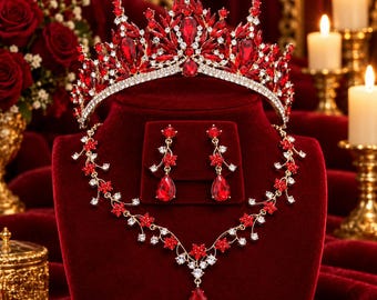 May include: A gold-tone jewelry set featuring a tiara, a necklace, and drop earrings. Each piece is adorned with deep red teardrop-shaped crystals and small clear rhinestones arranged in floral and vine-like patterns on a metal base.