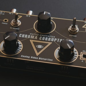 May include: A black rectangular analog video distortion device labeled Chroma Corruptor by Melted Electronics. It features two large black knobs, two toggle switches, two slide switches, and gold-colored printed graphics including an eye icon.