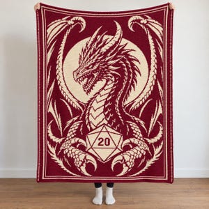 May include: A rectangular crochet blanket featuring a deep red and cream color scheme. The design displays a large, detailed dragon with spread wings centered behind a twenty-sided die graphic with the number 20 clearly visible in the center.