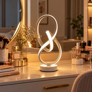 May include: A white LED table lamp featuring a continuous, intertwined loop design that emits a soft, warm glow. The lamp stands on a circular base and is connected to a power cord, showcasing a modern, sculptural form with smooth edges.