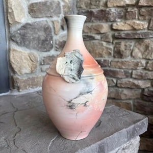 May include: A ceramic vase with a tapered neck and a rounded body, featuring a gradient of soft peach and cream tones with dark charcoal smoke-like markings. An embossed, textured gray leaf-shaped appliqué is attached to the upper side.