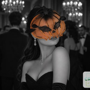 May include: An orange pumpkin-shaped masquerade mask featuring black mesh detailing, dark feathers, and black sequins. The mask is worn over the eyes, paired with a black strapless garment and a silver drop earring and necklace.