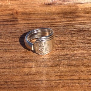 May include: A silver-toned ring crafted from a repurposed spoon handle. The band features a wrap-around design with a flat, rectangular face displaying an engraved geometric pattern consisting of small squares and lines on its surface.
