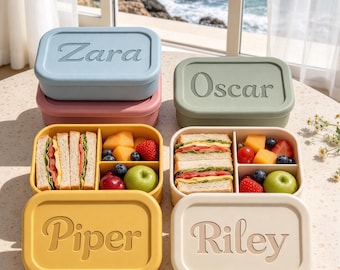 May include: Four rectangular silicone lunch boxes with lids featuring embossed names Zara, Oscar, Piper, and Riley. Two boxes are open, revealing compartments filled with sandwiches, melon chunks, strawberries, blueberries, and green apples.