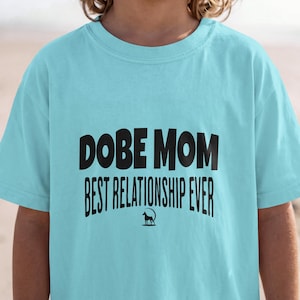 Op de afbeelding: Een lichtblauw t-shirt met zwarte tekst die DOBE MOM BEST RELATIONSHIP EVER leest, vergezeld door een kleine zwarte silhouetgrafiek van een Dobermann die in een dunne zwarte cirkelvormige omtrek onder de tekst staat.