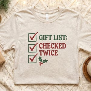 A lifestyle flat-lay of the heather beige short-sleeve checked it twice Christmas t-shirt showing the three-checkbox "GIFT LIST: CHECKED TWICE" design on a soft neutral background.