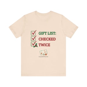 A cream mockup of a short-sleeve t-shirt with the checked it twice Christmas design showing three green checkboxes, the text "GIFT LIST: CHECKED TWICE," and a small open planner illustration.