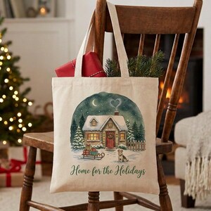 May include: A cream-colored canvas tote bag featuring a printed illustration of a snow-covered cottage at night with a dog, a sled with gifts, and the text Home for the Holidays in green script lettering centered on the front of the bag.