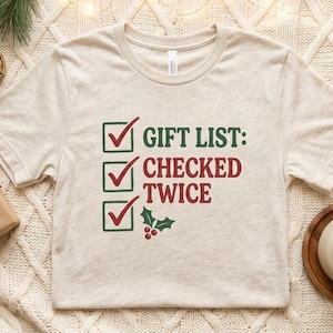 May include: A heather beige short-sleeve t-shirt featuring a graphic of three green check boxes with red check marks. The text reads "GIFT LIST: CHECKED TWICE" in green and red lettering, accompanied by a small green holly and red berry icon.
