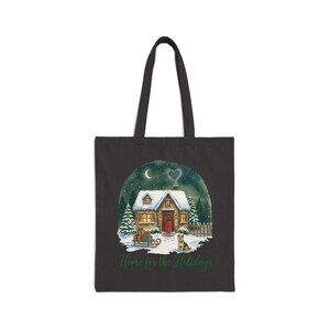 A black canvas tote bag in the Black color variant showing the snowy Christmas cottage "Home for the Holidays" print with a heart-shaped smoke plume on a plain light-gray background.