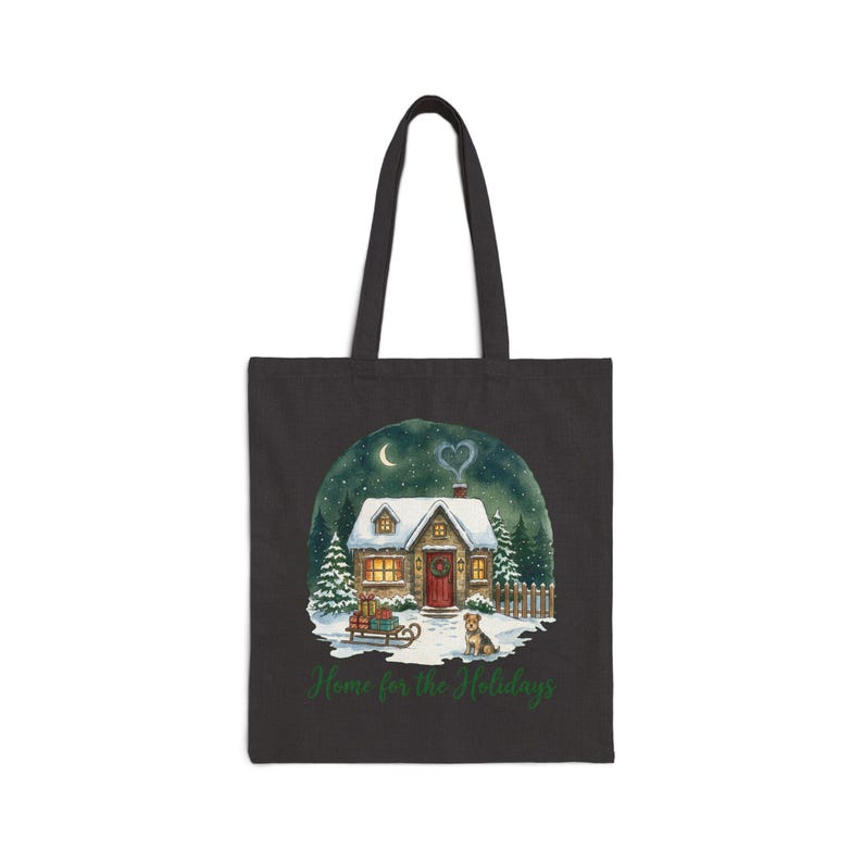 A black canvas tote bag in the Black color variant showing the snowy Christmas cottage "Home for the Holidays" print with a heart-shaped smoke plume on a plain light-gray background.