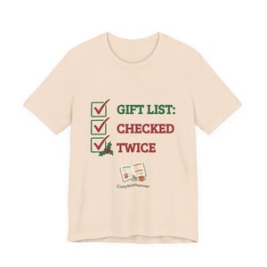 A second cream mockup angle of the short-sleeve checked it twice Christmas t-shirt with the three-checkbox "GIFT LIST: CHECKED TWICE" design and a small open planner illustration.