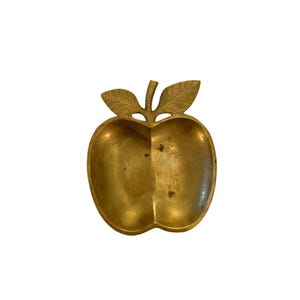 May include: A gold-toned metal dish shaped like an apple with a central stem and two leaves. The surface of the dish has a smooth, polished finish with subtle variations in color and texture, featuring a deep, rounded basin for holding small items.