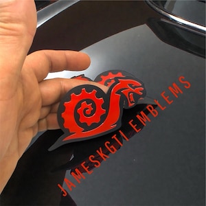 May include: A black and red snail-shaped emblem with a gear-like shell design. The emblem features a stylised snail with a sharp, aggressive head and a serrated shell. The text JAMESKGTI EMBLEMS is printed in red on the black surface.