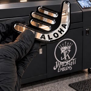 May include: A reflective, shaka-hand-shaped emblem with a black and silver finish. The word ALOHA is printed in bold, black capital letters across the center of the hand gesture. The emblem has a smooth, glossy surface and a distinct, raised border.