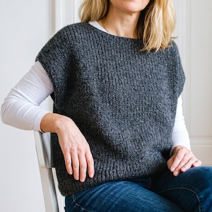 May include: A dark charcoal gray sleeveless knit sweater vest with a wide round neckline and a thick ribbed hem, worn over a long-sleeved white shirt and blue denim jeans. The sweater features a chunky, textured wool-blend knit pattern.