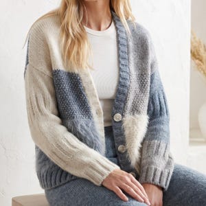 May include: A color-blocked cardigan featuring a mix of cream, light blue, and grey panels. The knit garment has a V-neckline, long sleeves with ribbed cuffs, and a front button closure with four round buttons. The texture varies between smooth knit, ribbed sections, and fuzzy patches.