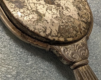 May include: A handheld mirror with a tarnished silver-tone finish and a decorative floral pattern on the back. The handle features vertical ridges and ornate scrollwork at the base where it connects to the oval mirror frame.