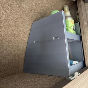 May include: A light blue, two-tiered plastic storage organizer with an angled front profile. The unit features an open-front design on both levels, holding a green-labeled spray bottle on the top shelf and a small tube on the bottom shelf.