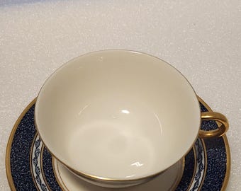 May include: A white porcelain teacup and saucer set featuring a dark blue speckled border with a delicate white vine pattern. The rim of the cup and the edges of the saucer are trimmed in gold, and the cup includes a gold-colored handle.