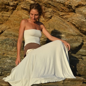 May include: A woman wearing a white strapless top and a long white skirt sits on a rocky cliff. She has long brown hair and is wearing earrings.