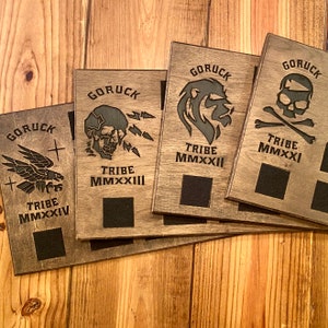 May include: Four wooden plaques with the word "GORUCK" and the text "TRIBE" followed by a Roman numeral. Each plaque features a different graphic: a skull and crossbones, a lion, a skull with lightning bolts, and a bird with wings spread.