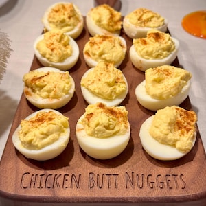 May include: A wooden serving board with deviled eggs. The eggs are cut in half, filled with yellow filling, and sprinkled with paprika. The board is engraved with the words "CHICKEN BUTT NUGGETS".