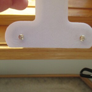 May include: A pair of small, round, silver-toned stud earrings featuring a single clear, faceted stone in the center of each. The earrings are mounted on a light purple, T-shaped foam display card designed for holding jewelry securely in place.