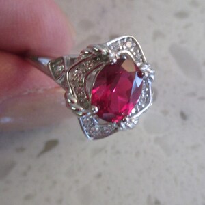 May include: A silver-toned ring featuring a large, oval-cut, deep red gemstone at the center. The stone is surrounded by a rectangular frame embellished with small, clear, sparkling stones. The band has a polished finish and a tapered design.